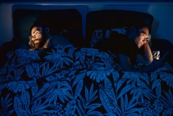 couple in bed under the sheets turning back on each other and looking at phones