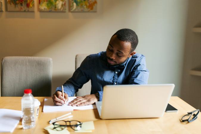man with laptop writing in notebook