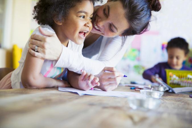 Affectionate, happy mother hugging toddler daughter coloring at table