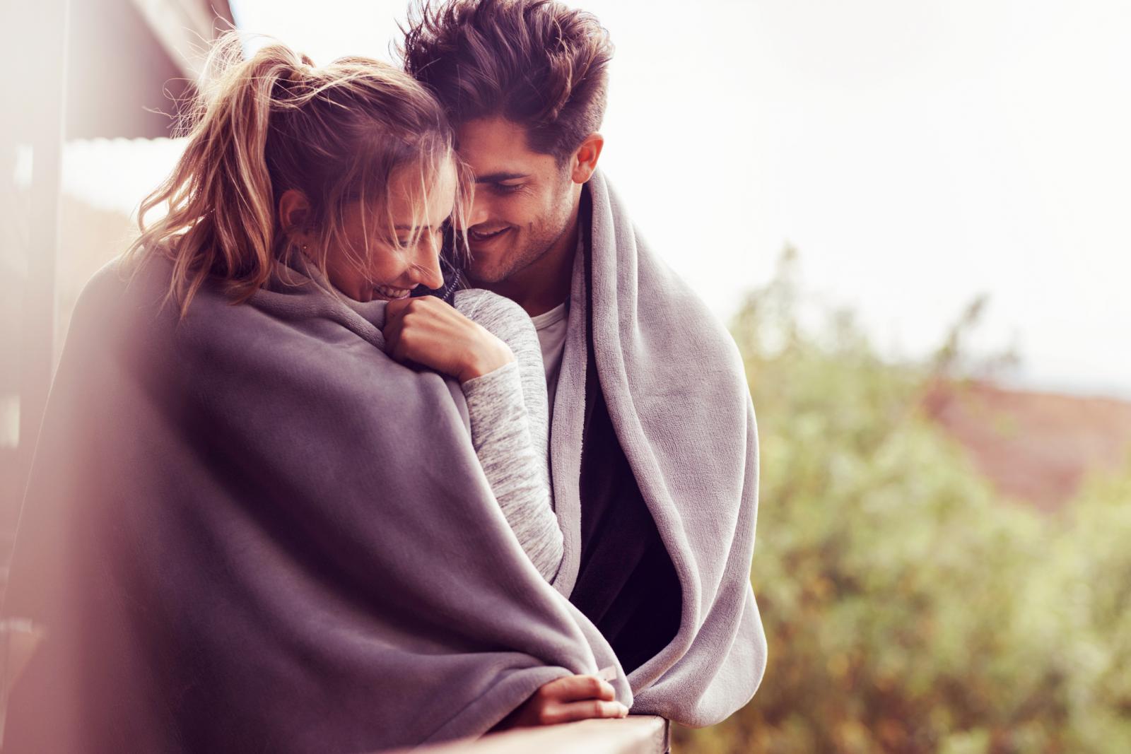 Man and woman standing together in a balcony wrapped in a blanket embracing and smiling