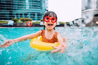 Playful little girl with sunglasses smiling joyfully, splashing and playing with water happily in the swimming pool on Summer vacations