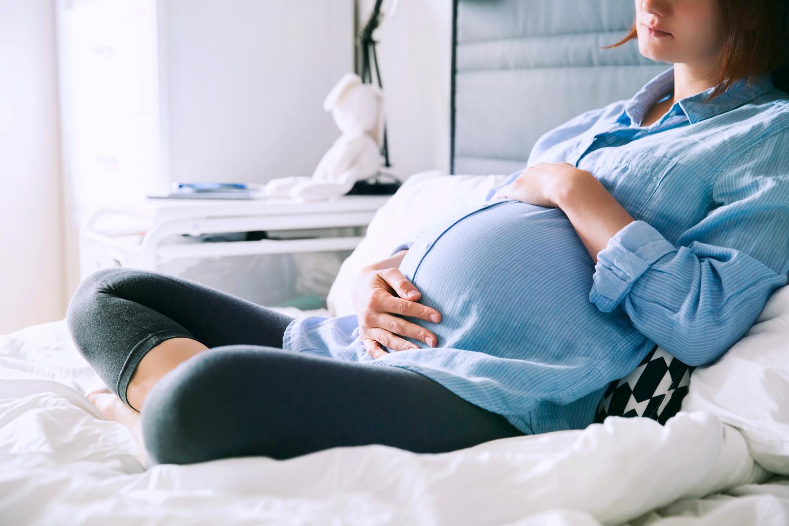 Pregnant woman sitting at bed and holds hands on belly in bedroom at home